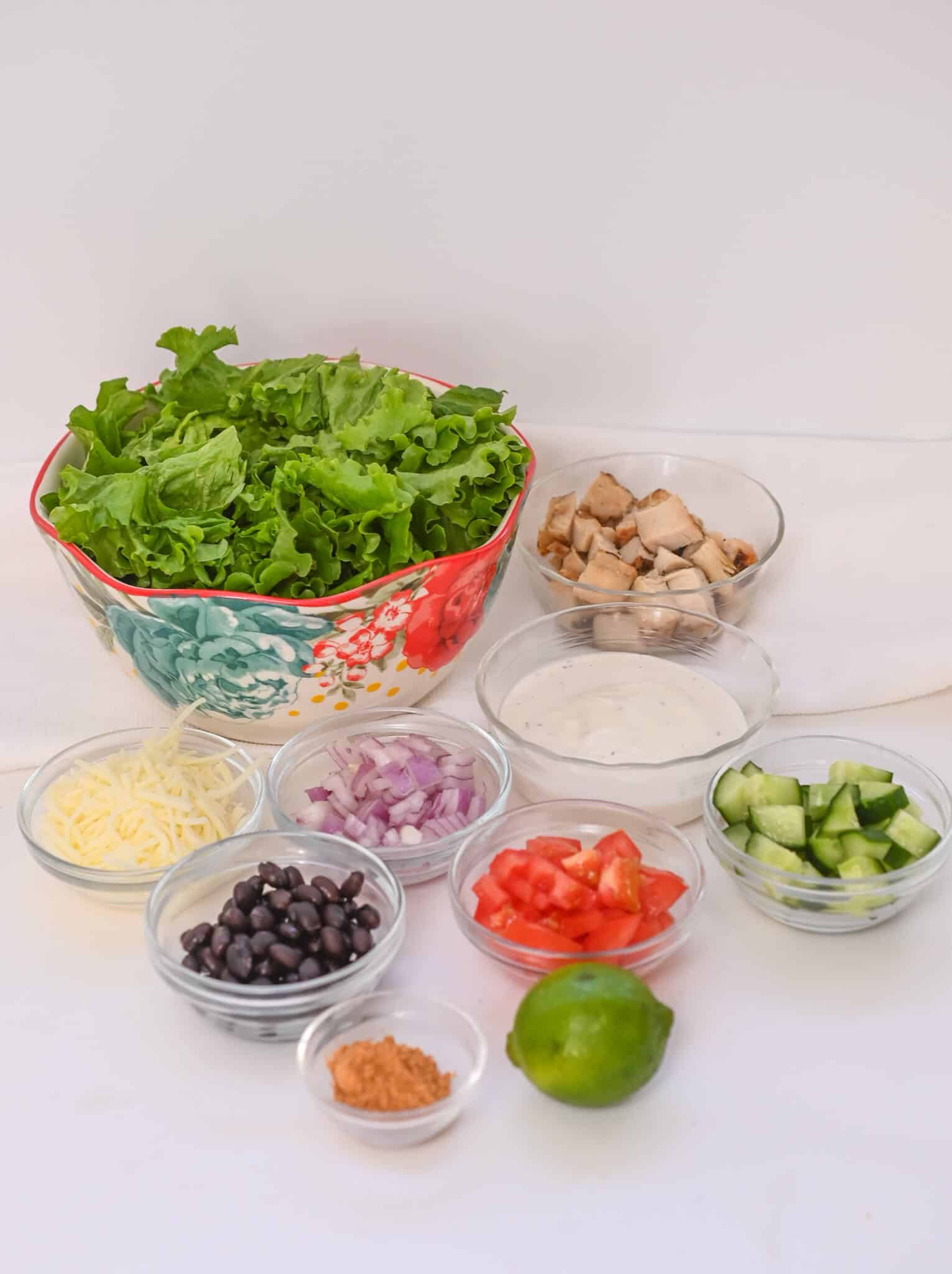 A bowl of lettuce with small bowls of chopped chicken, shredded cheese, black beans, onions, tomatoes, cucumbers, dressing, taco seasoning, and a lime arranged around it.