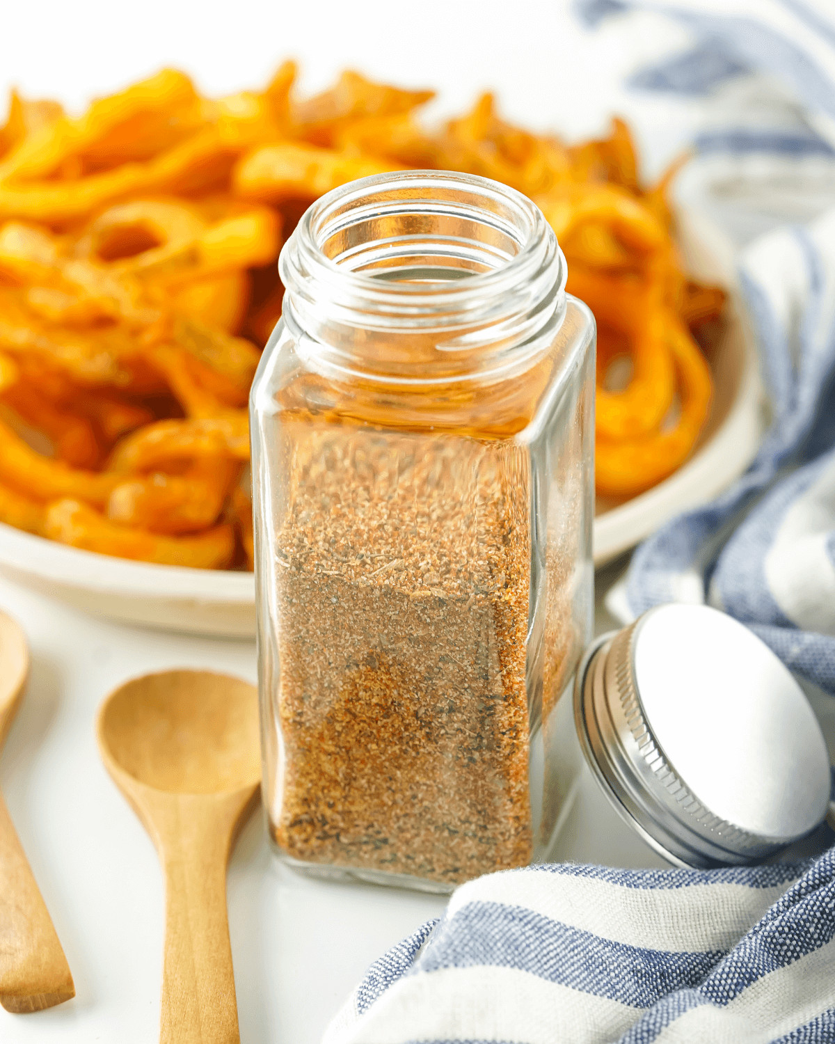 A glass jar of French fry seasoning next to a wooden spoon.