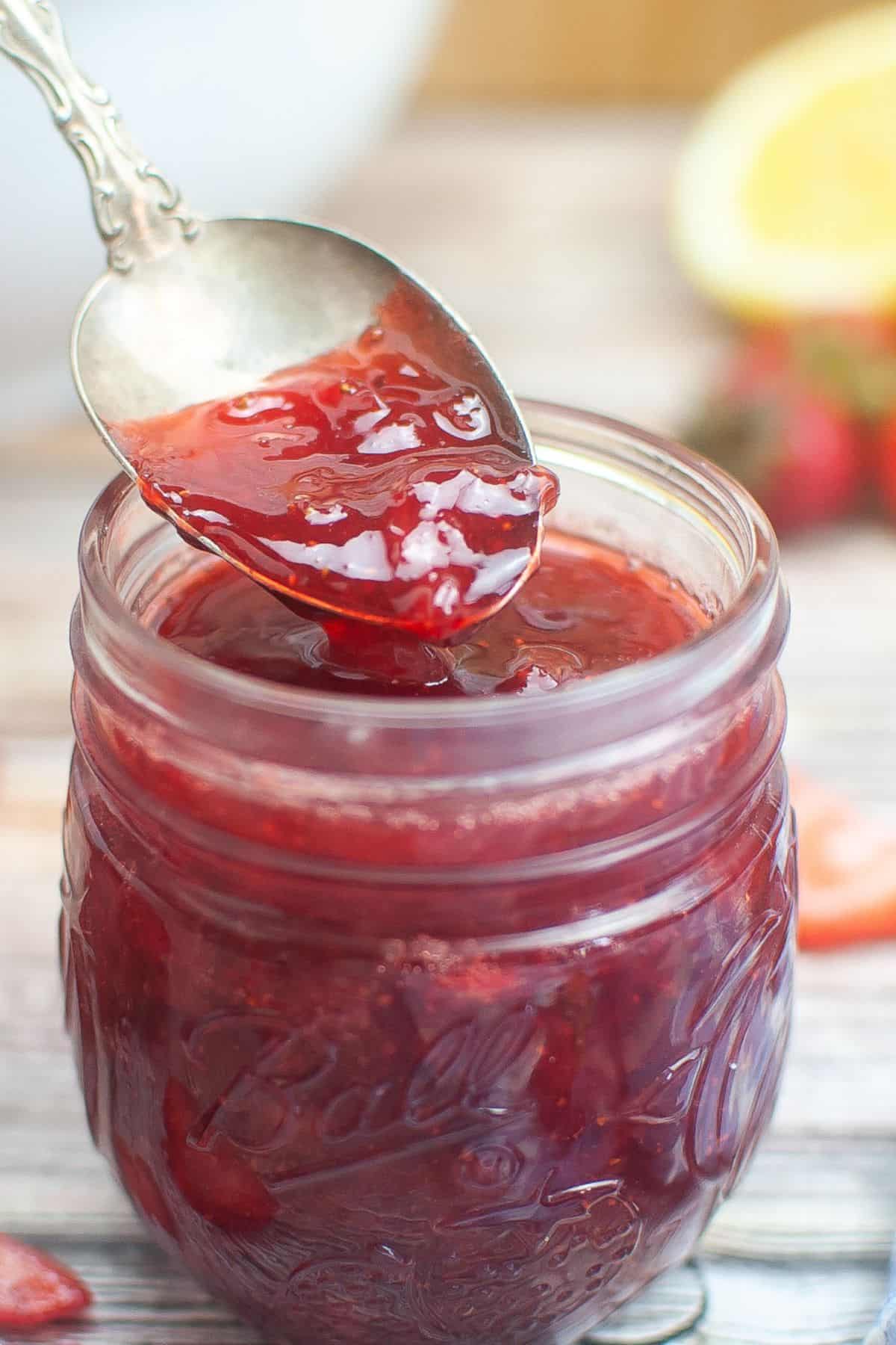 A spoon holding No Pectin Strawberry Jam above a glass jar filled with jam, with sliced strawberries and a lemon slice in the background.