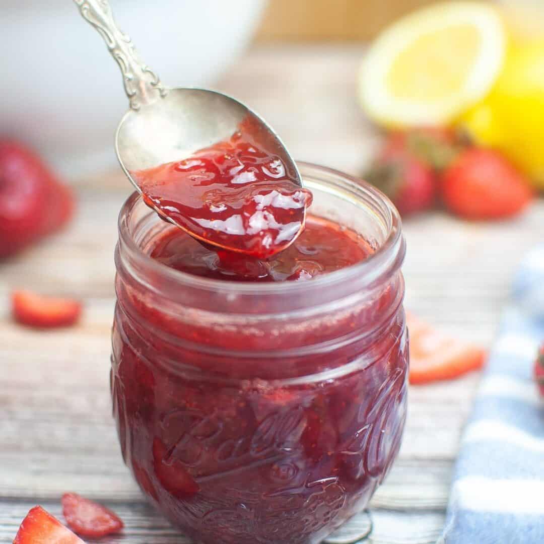 A spoon holds No Pectin Strawberry Jam above a glass jar brimming with jam, with sliced strawberries and a lemon in the background.