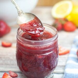 A spoon holds No Pectin Strawberry Jam above a glass jar brimming with jam, with sliced strawberries and a lemon in the background.