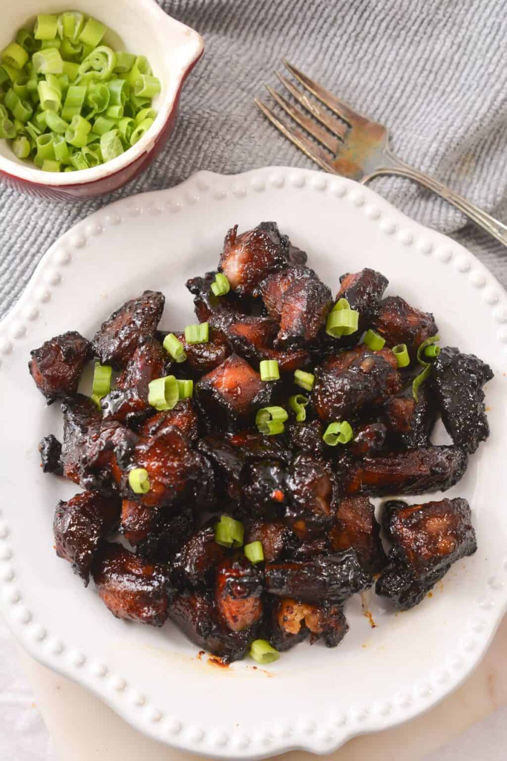 A white plate with glazed, dark brown Pork Burnt Ends topped with chopped green onions, next to a small bowl of extra green onions and a fork.