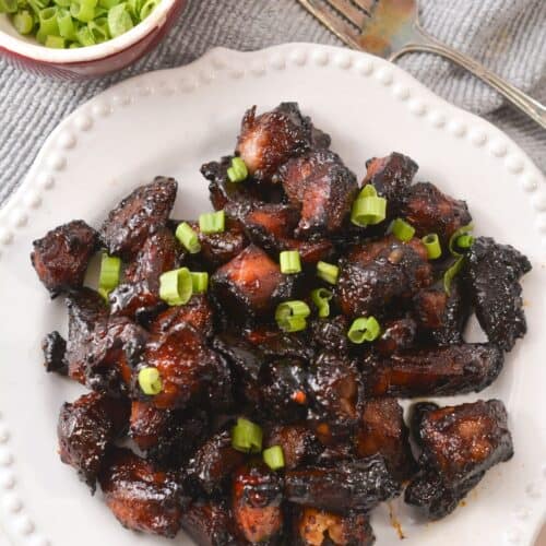 A white plate with glazed, dark brown Pork Burnt Ends topped with chopped green onions, next to a small bowl of extra green onions and a fork.