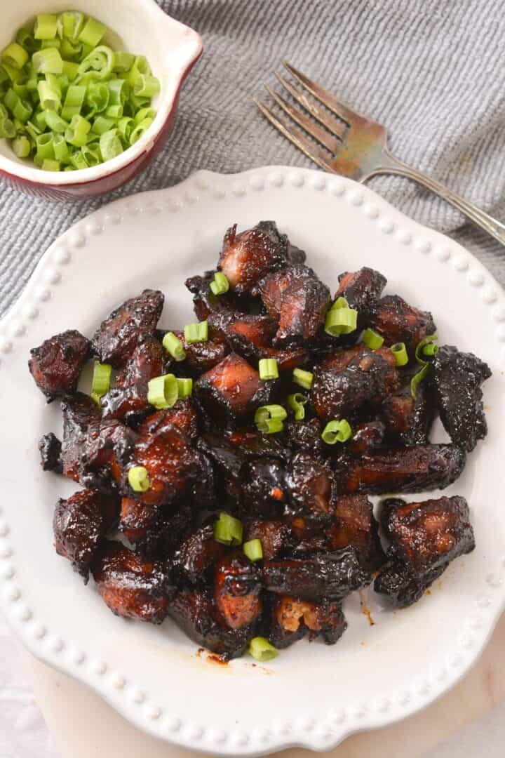 A white plate with glazed, dark brown Pork Burnt Ends topped with chopped green onions, next to a small bowl of extra green onions and a fork.