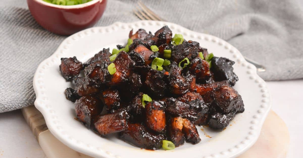 A plate of glazed, dark-colored Pork Burnt Ends garnished with chopped green onions, served on a white plate with a fork and a bowl of more garnish in the background.