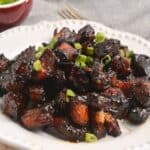 A plate of glazed, dark-brown Pork Burnt Ends garnished with chopped green onions, served on a white dish with a bowl of extra green onions in the background.
