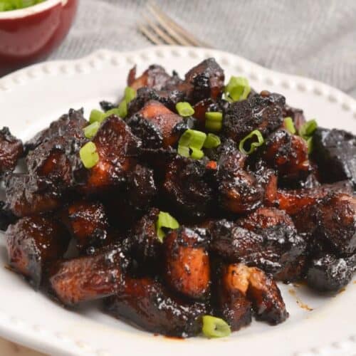 A plate of glazed, dark-brown Pork Burnt Ends garnished with chopped green onions, served on a white dish with a bowl of extra green onions in the background.
