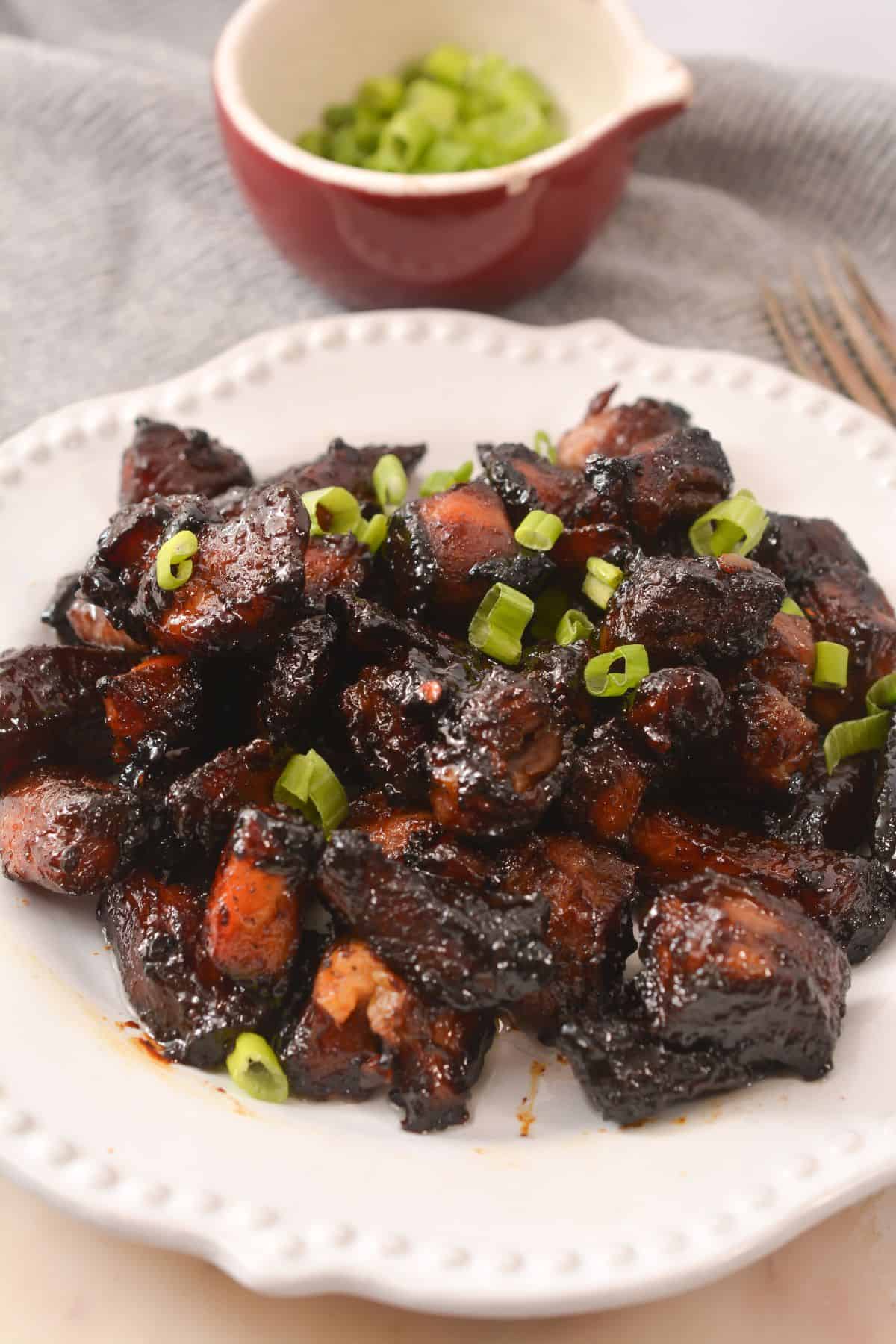 A white plate with glazed, browned chunks of pork burnt ends garnished with chopped green onions; a bowl of additional green onions is in the background.