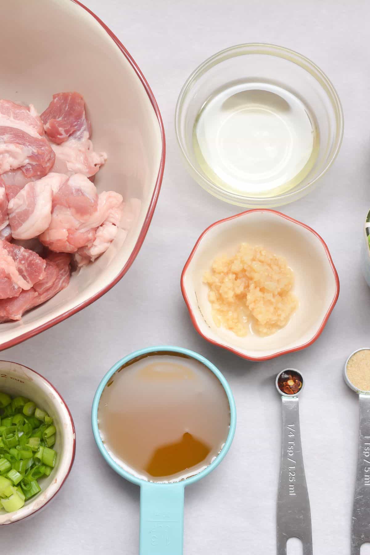 Raw pork pieces for Pork Burnt Ends, chopped green onions, minced garlic, a measuring cup of broth, oil in a glass bowl, and measuring spoons with spices arranged on a white surface.