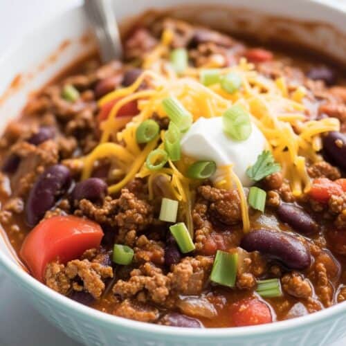Bowl of venison chili with ground meat, kidney beans, tomatoes, topped with shredded cheese, sour cream, and chopped green onions. A spoon is in the bowl—perfect for trying out your favorite venison chili recipe.