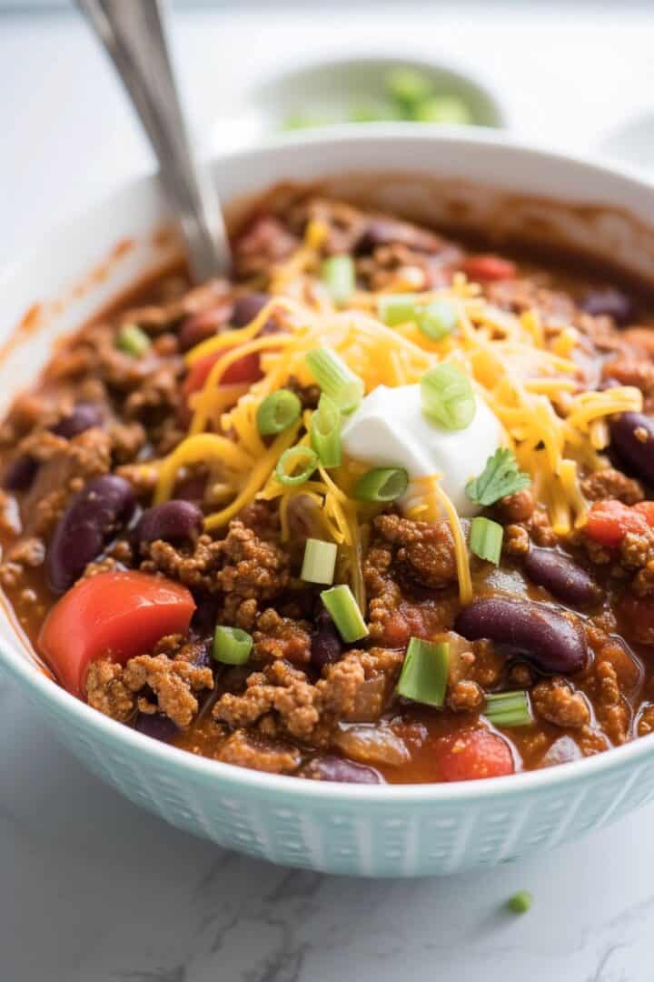 Bowl of venison chili with ground meat, kidney beans, tomatoes, topped with shredded cheese, sour cream, and chopped green onions. A spoon is in the bowl—perfect for trying out your favorite venison chili recipe.