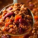 A close-up of a spoon holding Venison Chili with ground beef, kidney beans, tomato sauce, and diced carrots over a pot of the same chili.
