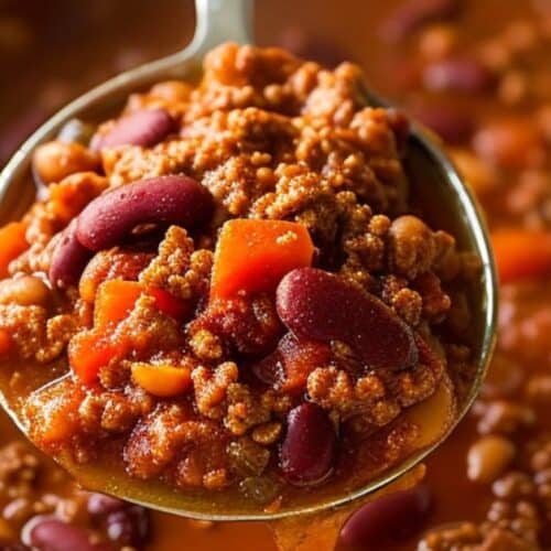 A close-up of a spoon holding Venison Chili with ground beef, kidney beans, tomato sauce, and diced carrots over a pot of the same chili.
