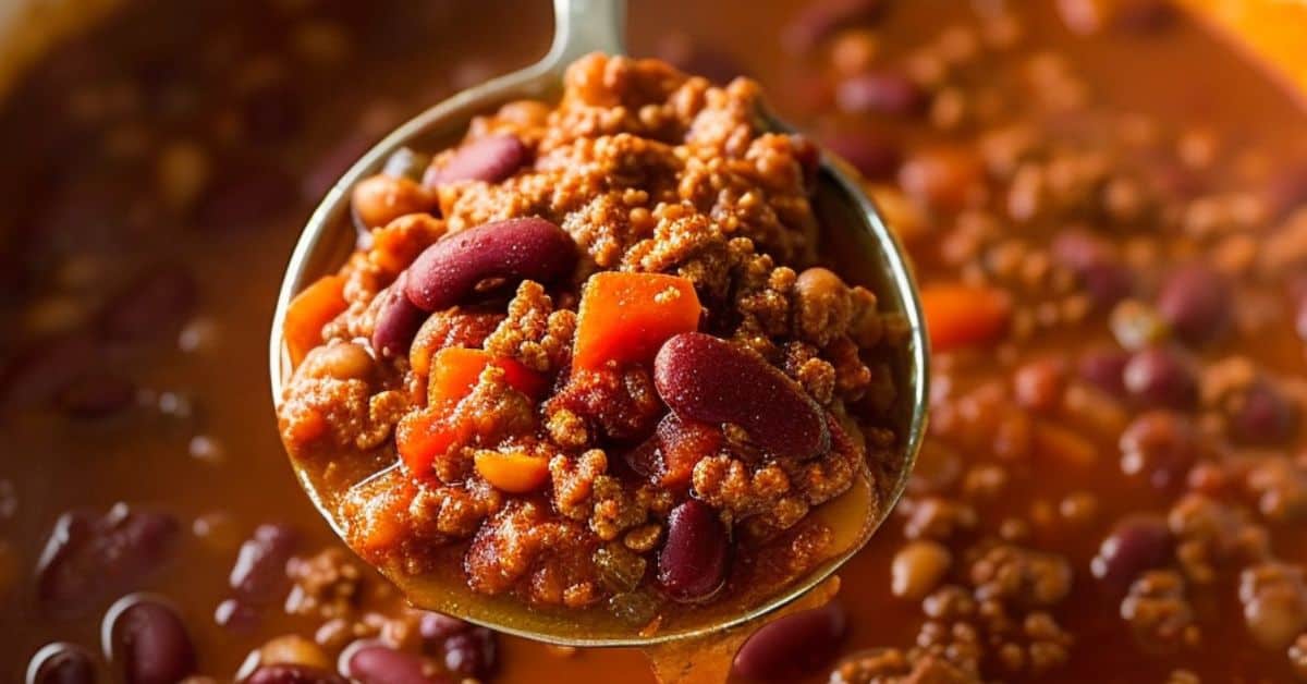 A metal ladle holds Venison Chili with ground meat, kidney beans, diced tomatoes, and carrots over a pot of the same chili.