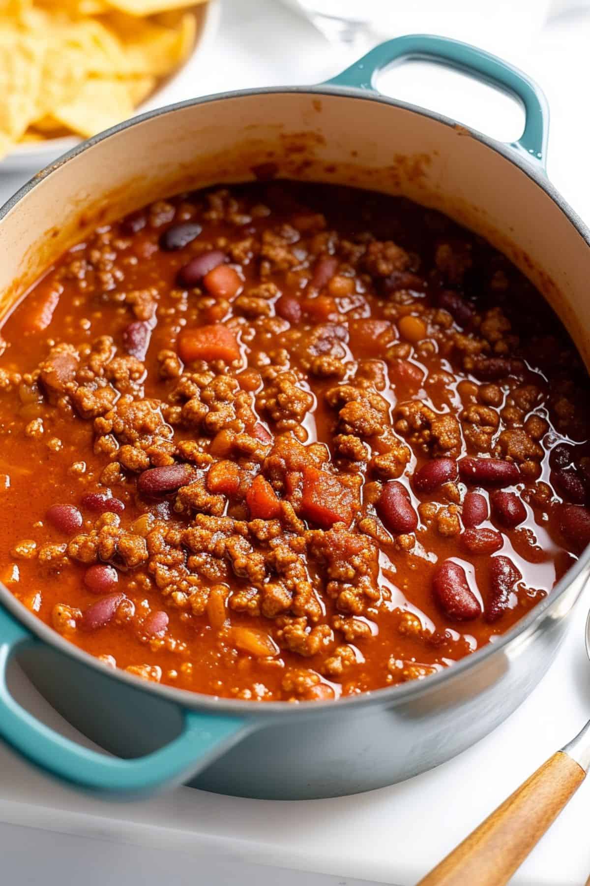 A pot of Venison Chili with ground beef, kidney beans, and tomato sauce is simmering on a stovetop, with tortilla chips visible in the background.