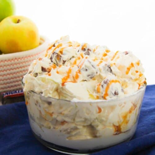 A glass bowl of creamy dessert salad with grapes and orange drizzle sits on a blue cloth. Apples, perfect for an easy recipe like Apple Snickers Salad, are in a basket in the background.