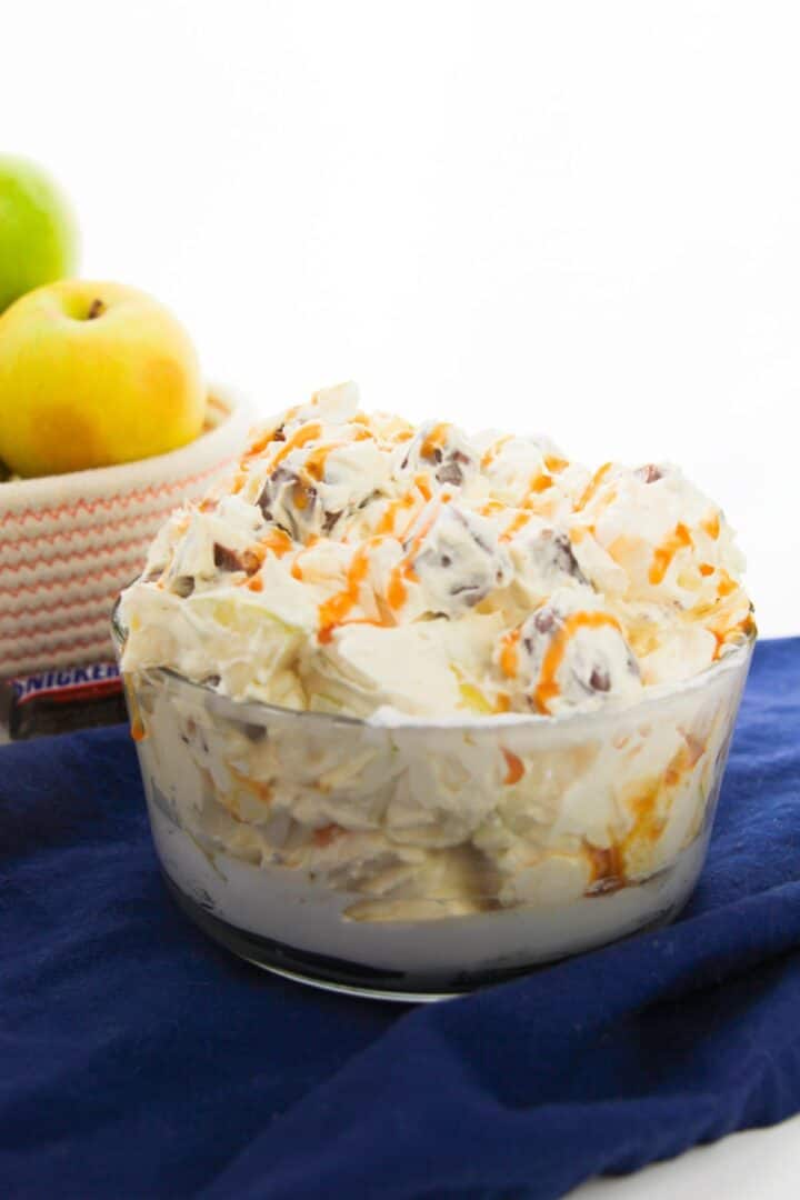 A glass bowl of creamy dessert salad with grapes and orange drizzle sits on a blue cloth. Apples, perfect for an easy recipe like Apple Snickers Salad, are in a basket in the background.