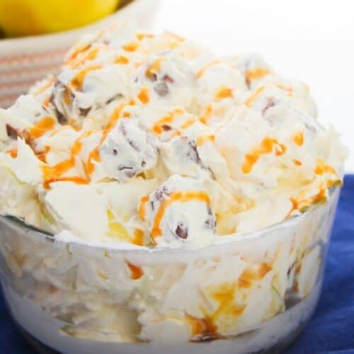 A glass bowl filled with creamy dessert salad, topped with caramel drizzle, sits on a blue cloth. A bowl of lemons is in the background, enhancing the inviting look of this Apple Snickers Salad-inspired apple dessert.