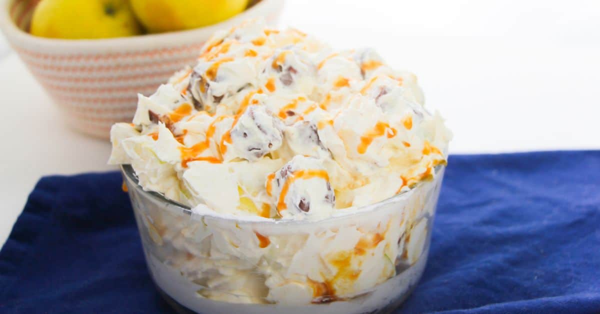 A glass bowl filled with creamy dessert salad, topped with caramel drizzle, sits on a blue cloth. A bowl of lemons is in the background, enhancing the inviting look of this Apple Snickers Salad-inspired apple dessert.