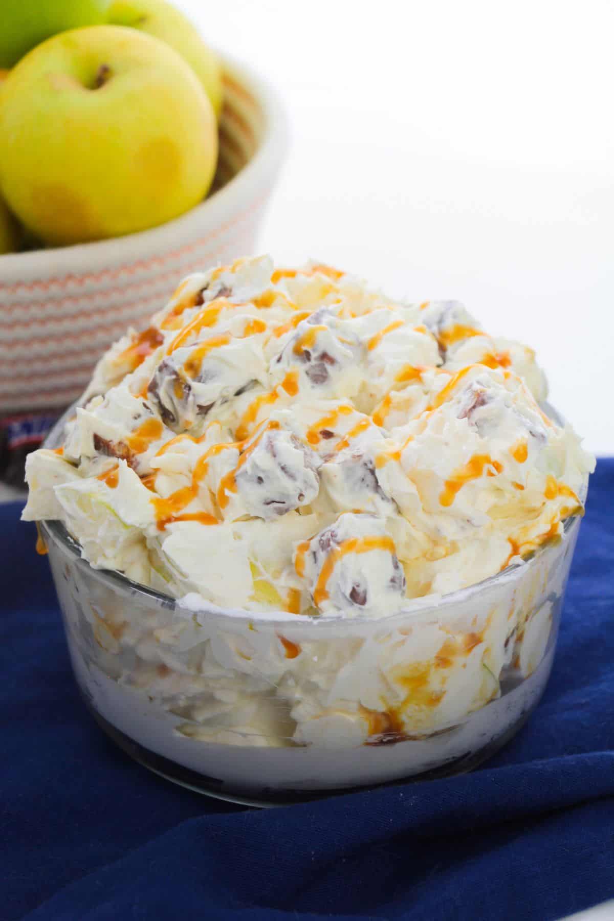 A glass bowl filled with creamy Apple Snickers Salad, topped with drizzled caramel, sits on a blue cloth. A basket of apples is in the background.
