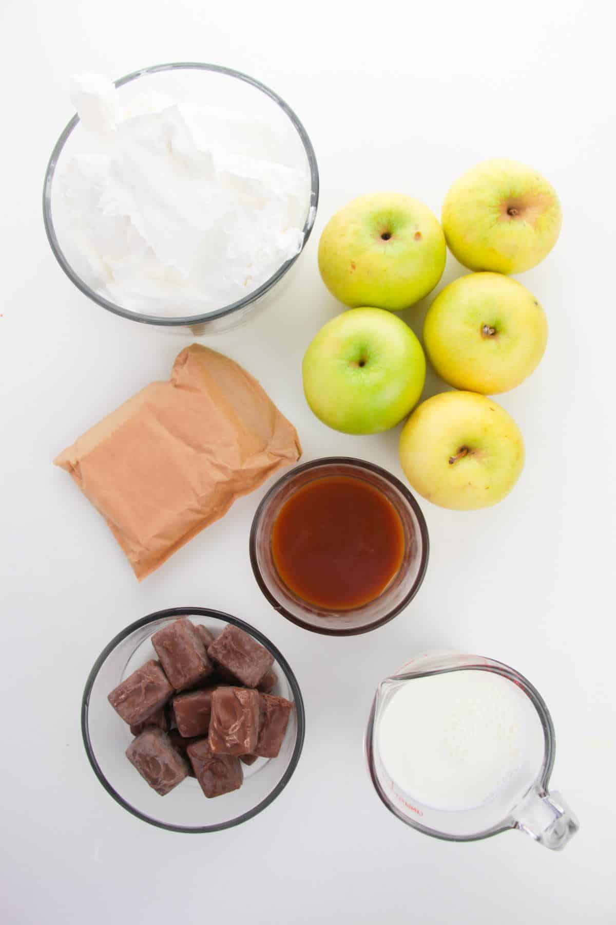 Six green apples, a glass bowl of whipped topping, wrapped caramels, a brown packet, a glass of milk, and a bowl of caramel sauce are arranged on a white background—perfect ingredients for creating a delightful Apple Snickers Salad dessert salad.