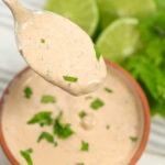 A spoonful of creamy Baja Chipotle sauce with chopped herbs, above a bowl, with fresh lime and lettuce in the background.