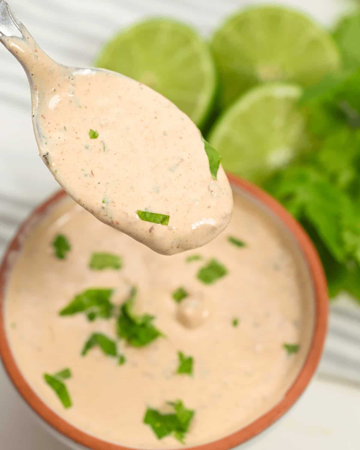 A spoonful of creamy Baja Chipotle sauce with chopped herbs, above a bowl, with fresh lime and lettuce in the background.