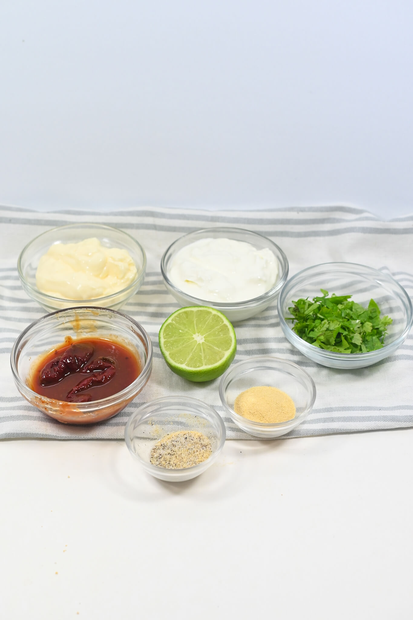 Assorted condiments and seasonings in small bowls, including mayonnaise, lime, cream, chopped herbs, spices, and Baja chipotle sauce.
