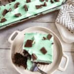 A plate with a slice of chocolate mint cake topped with mint green frosting and chocolate pieces, with more cake in the background.
