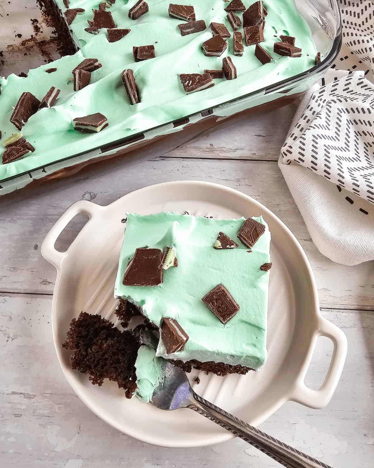 A plate with a slice of chocolate mint cake topped with mint green frosting and chocolate pieces, with more cake in the background.