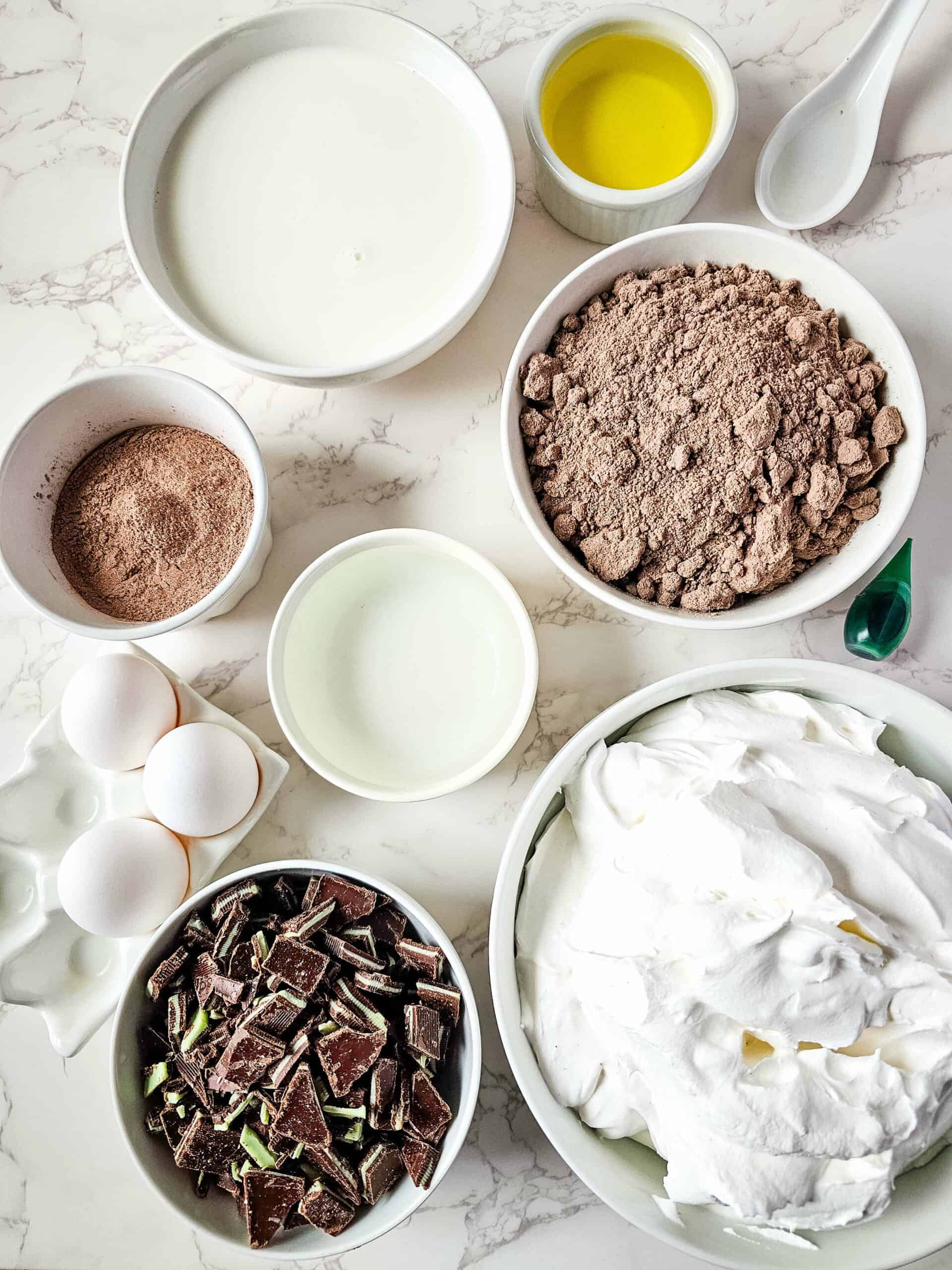 Ingredients for baking arrayed on a marble countertop, including bowls of flour, cocoa powder, eggs, sugar, chocolate mint chunks, and whipped cream.