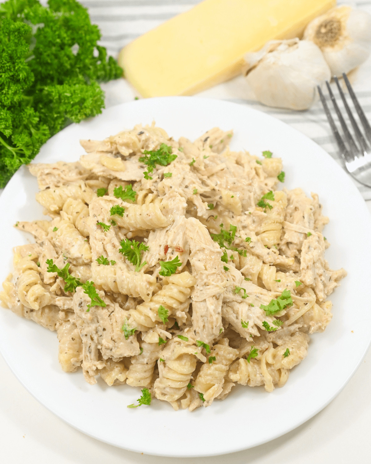 A plate of creamy crock pot garlic parmesan chicken pasta garnished with parsley, accompanied by cheese, garlic, and herbs in the background.
