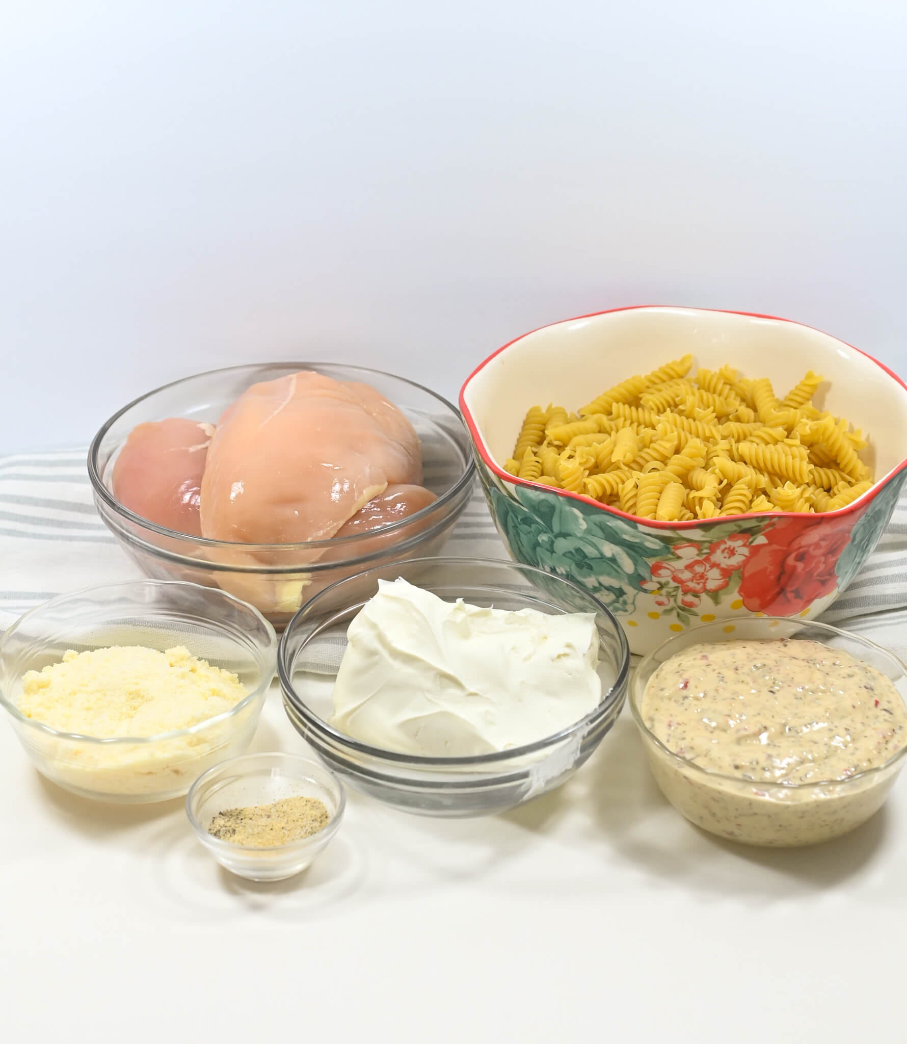 Ingredients for cooking laid out on a table, including raw chicken breasts, pasta, cream, spices, grated Parmesan cheese, and a bowl of garlic sauce.