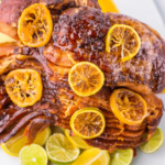 Meat with caramelized orange slices, accompanied by lime and orange wedges on a white plate.