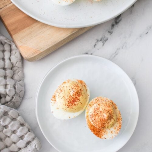 Two Deviled Eggs without Mayonnaise topped with paprika rest on a white plate, while more of these creamy, mayo-free deviled eggs are displayed on a serving platter in the background.