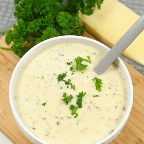 Bowl of creamy garlic parmesan sauce soup garnished with parsley on a wooden board, with cheese and more parsley in the background.