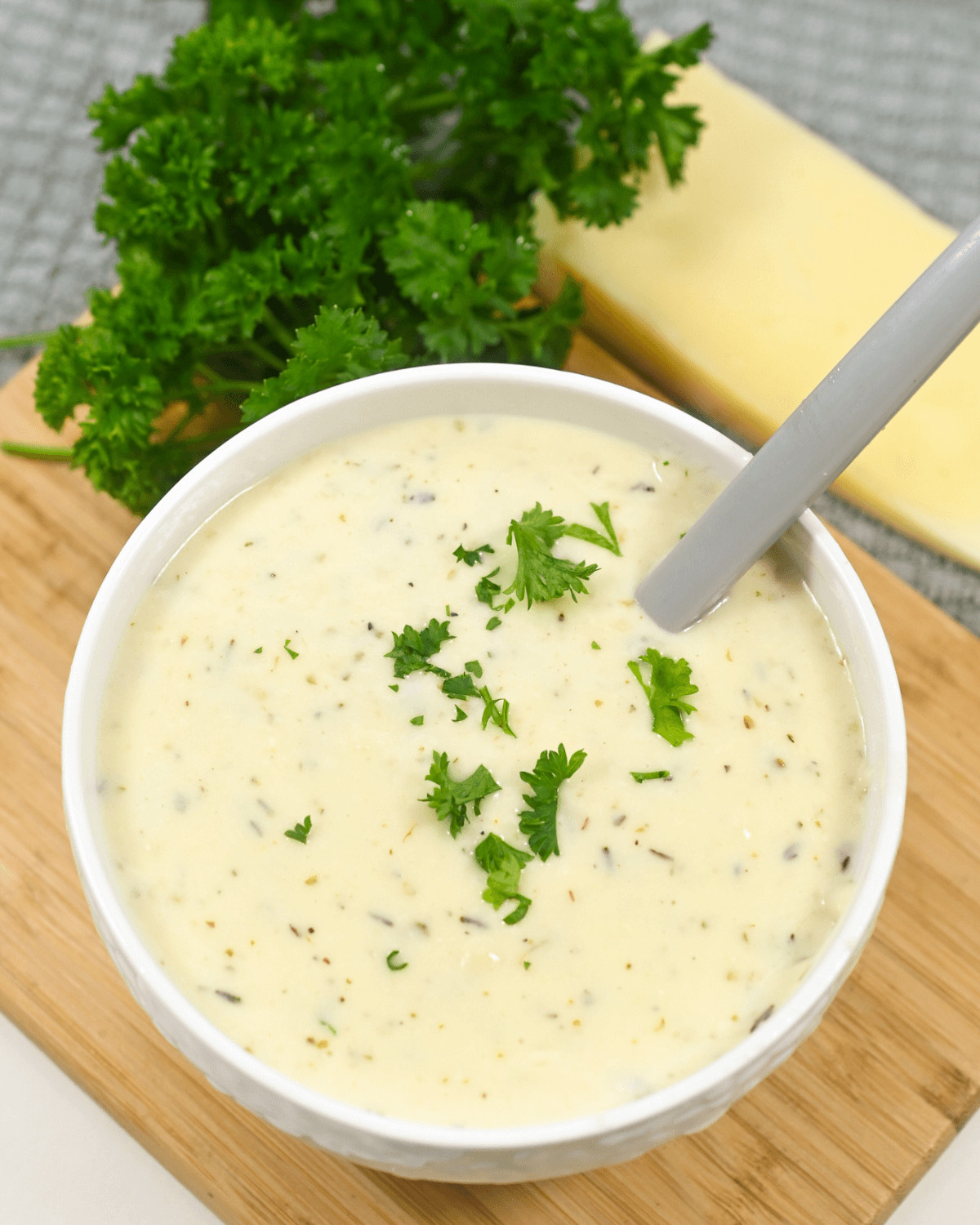 Bowl of creamy garlic parmesan sauce soup garnished with parsley on a wooden board, with cheese and more parsley in the background.