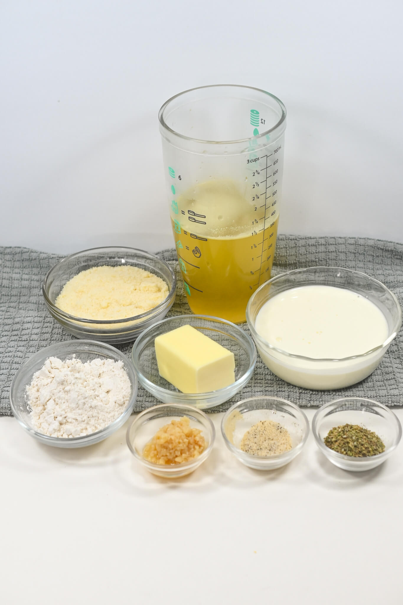 Several ingredients for baking arranged on a table, including flour, butter, garlic parmesan sauce, herbs, and liquids in glass containers.