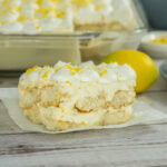 A slice of Lemon dessert with whipped cream and lemon zest on a white surface with a whole dessert in the background.