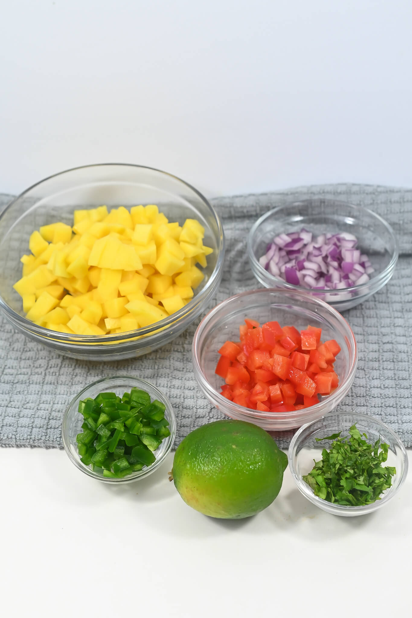 Prepped ingredients for a recipe, including diced mango, onions, tomatoes, jalapeños, lime, and cilantro in separate bowls for mango habanero salsa.