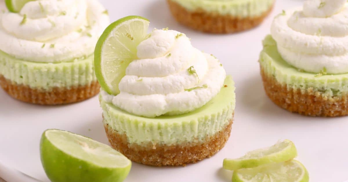 Mini Key Lime Cheesecake treats with graham cracker crusts, topped with whipped cream and a lime slice, are displayed on a white surface alongside extra lime slices.