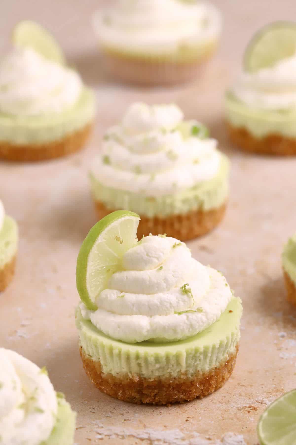 Mini Key Lime Cheesecake treats with a graham cracker crust, topped with whipped cream and a slice of lime, are artfully arranged on a light surface.