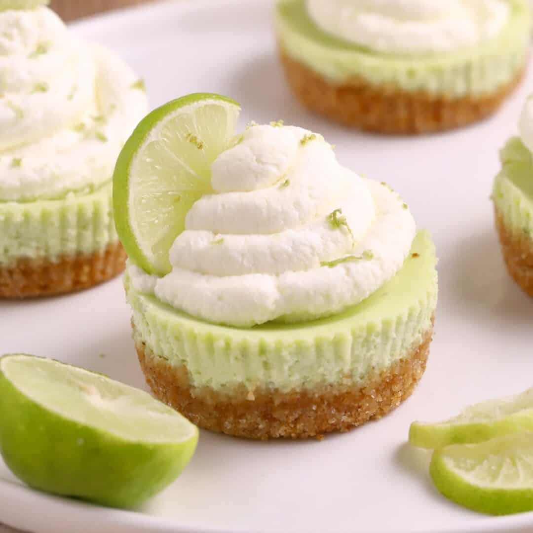 Mini Key Lime Cheesecake topped with whipped cream and a fresh lime slice, served on a white plate with extra lime slices for a zesty finish.