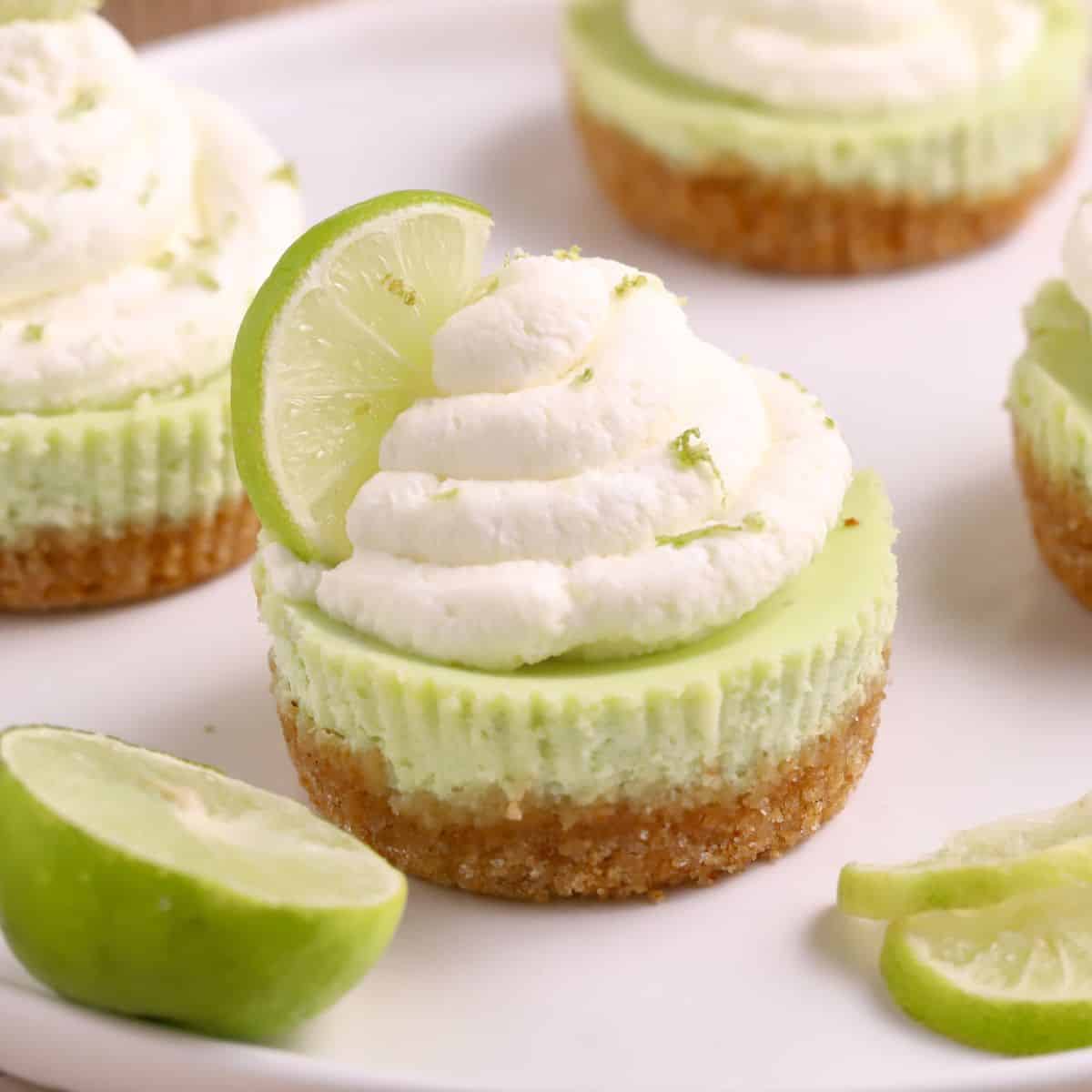 Mini Key Lime Cheesecake topped with whipped cream and a fresh lime slice, served on a white plate with extra lime slices for a zesty finish.