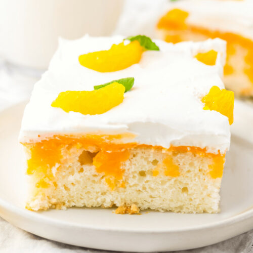 A slice of orange creamsicle cake with whipped cream topping and fruit garnish on a white plate.