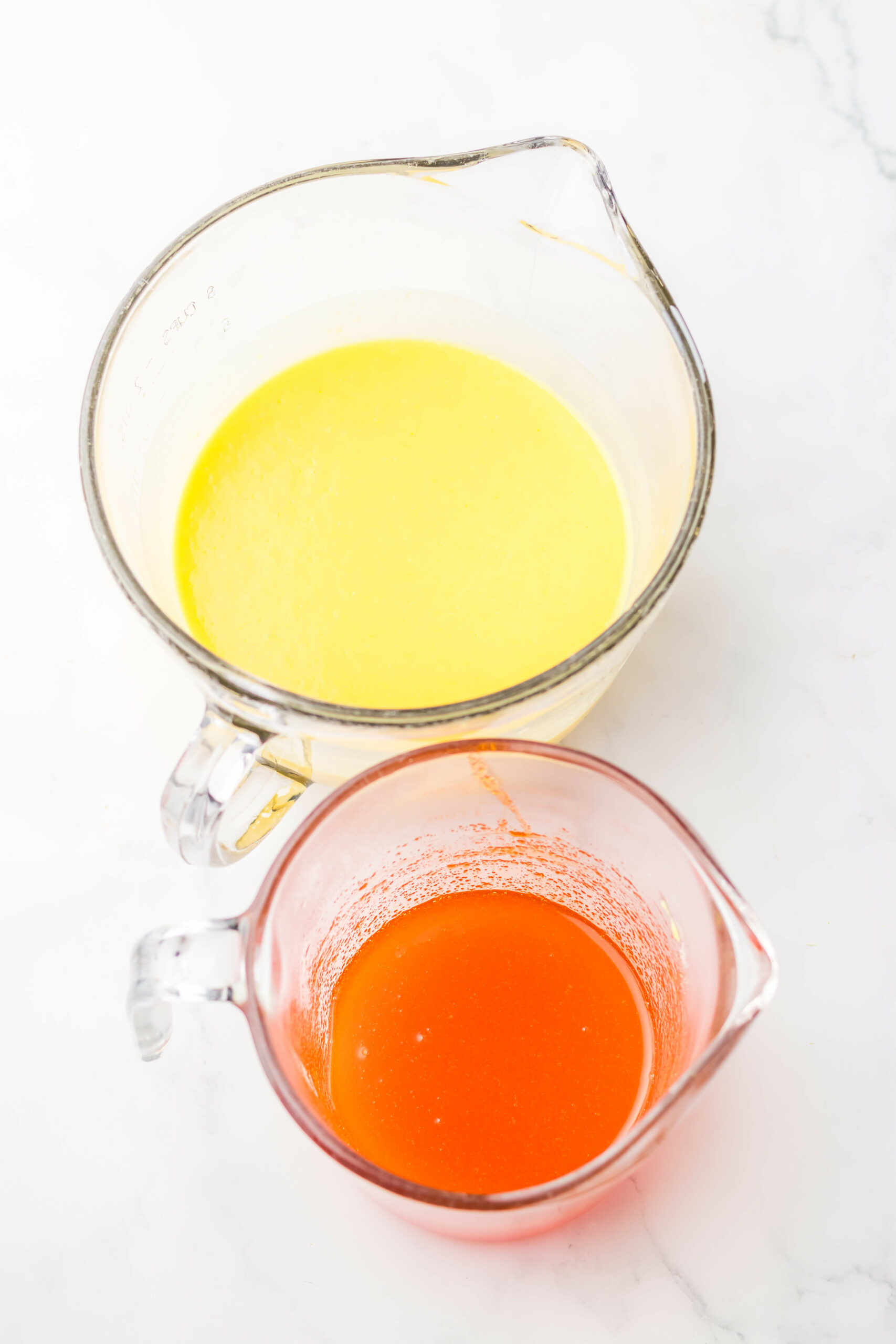 Two measuring jugs with liquid ingredients on a white surface — one contains yellow liquid, possibly melted butter, and the other contains red liquid for making an orange creamsicle cake.