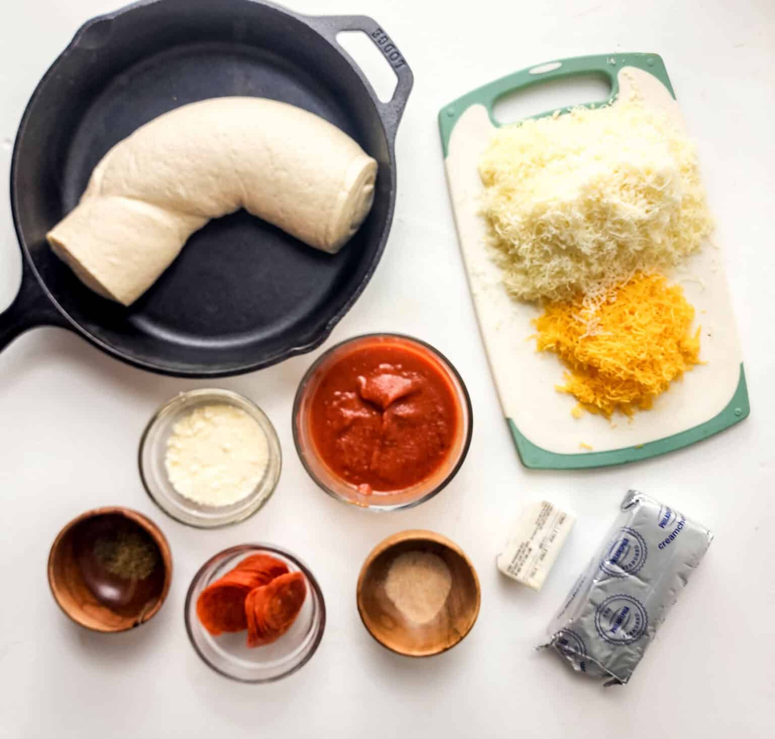 Ingredients for pizza dip arranged on a kitchen countertop, including dough, shredded cheese, sauce, and various seasonings.