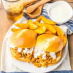 Two slow-cooker buffalo chicken sloppy joes with potato wedges on a plate, celery sticks on the side, and a glass of iced beverage.