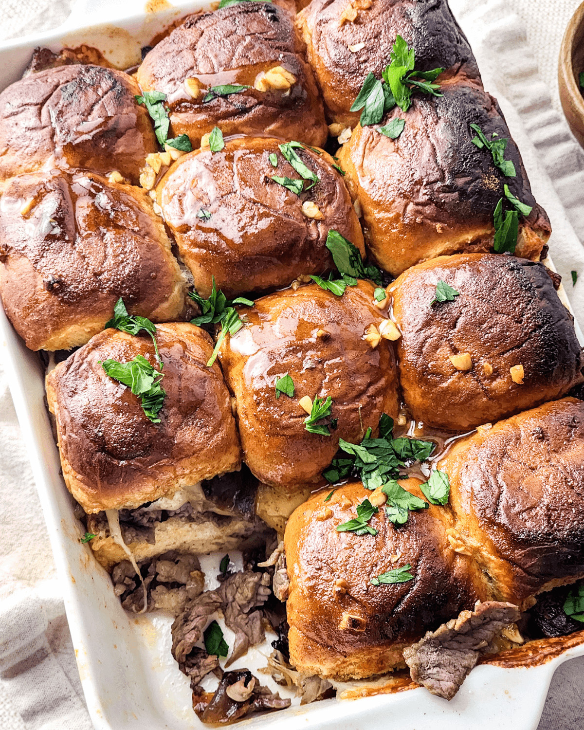 Baked steak and cheese sliders garnished with fresh parsley in a white baking dish.