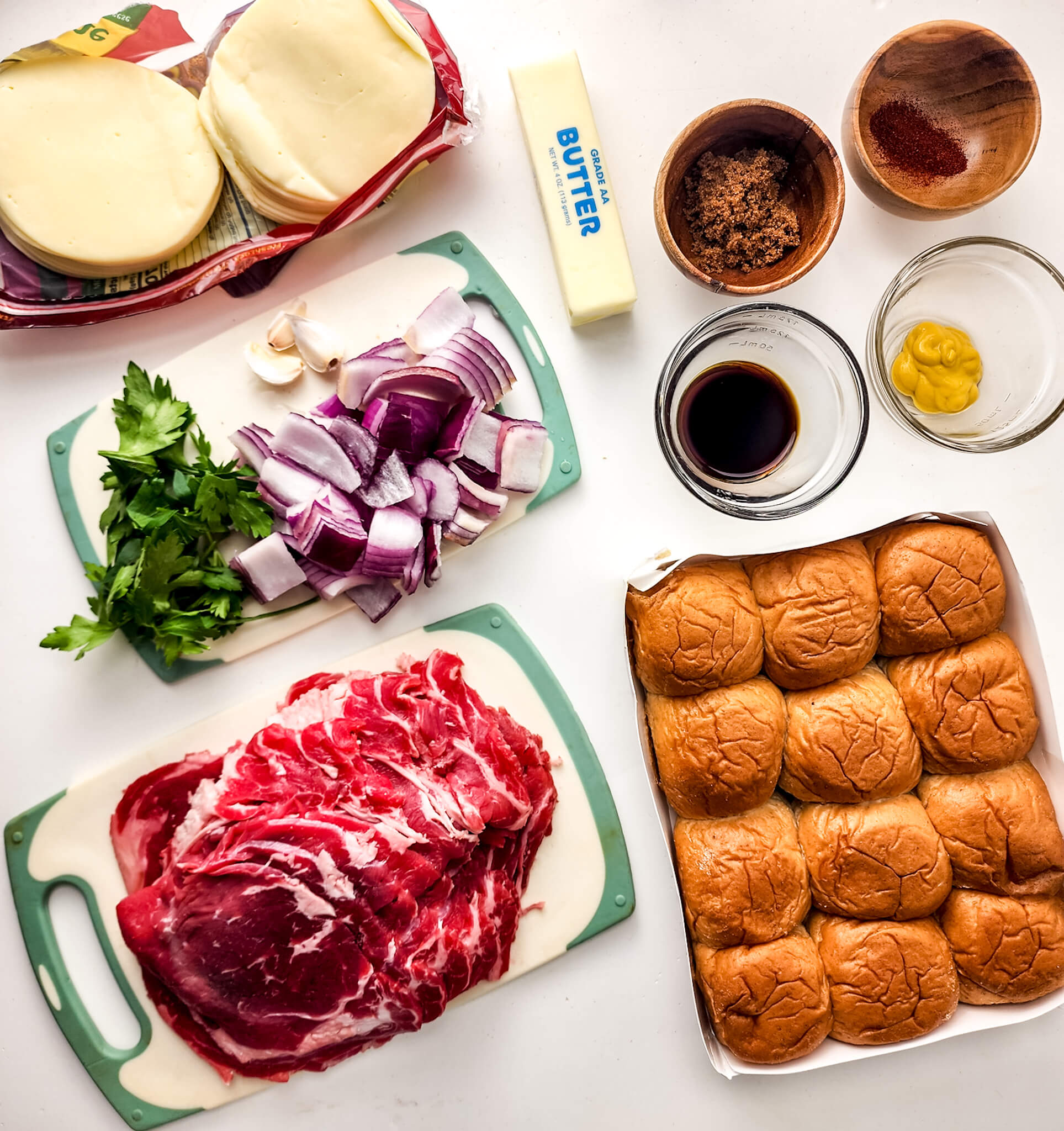Ingredients for steak and cheese sliders laid out on a counter, including steak, buns, cheese, butter, spices, and condiments.